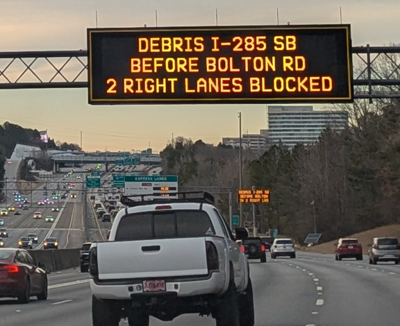 A white pickup truck drives along a highway with light traffic and oncoming vehicles visible across the median.